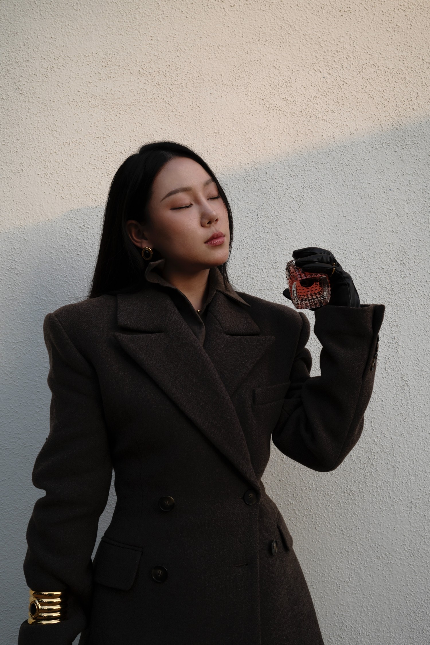 Brown tailored blazer with fragrance held in black leather gloves against a sunlit wall