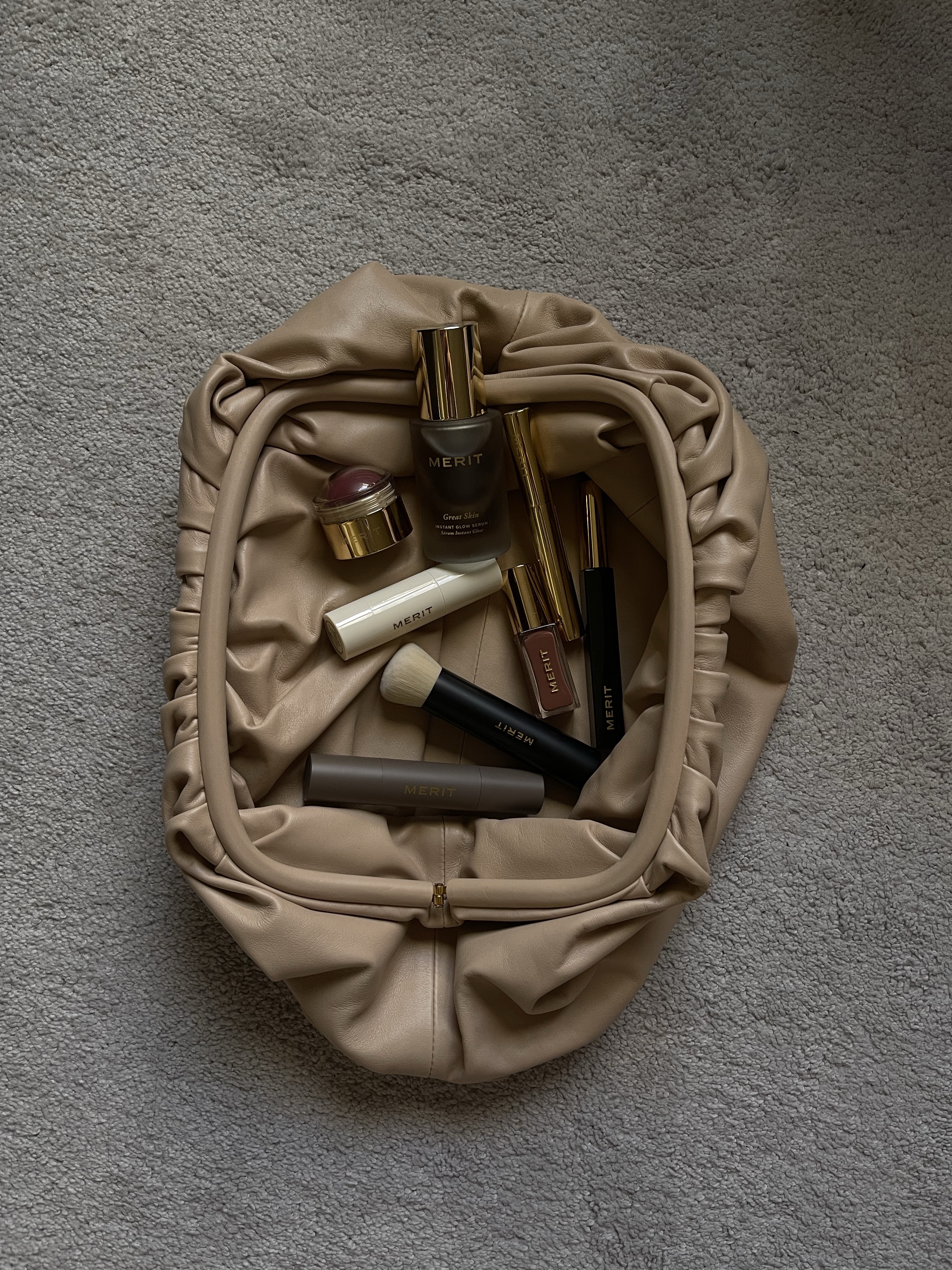 Merit beauty products spilling from a nude leather pouch on grey carpet